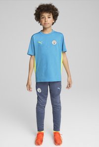 Vaaleansininen jalkapallosharja, jossa on keltaisia yksityiskohtia ja kuviomainen pinta; harmaat housut logolla; oranssit nappulakengät kirkkaaraisilla keltaisilla sukilla.