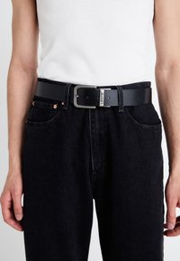 Zwarte leren riem met een zilveren metalen gesp. De gesp heeft de merknaam "Levi's." Draag over zwarte denim jeans.