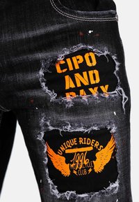 Jeans in denim nero con toppe strappate con testo arancione. I design più notevoli includono "CIPO AND RAYX" e "UNIQUE RIDERS 1996" con grafiche di ali.
