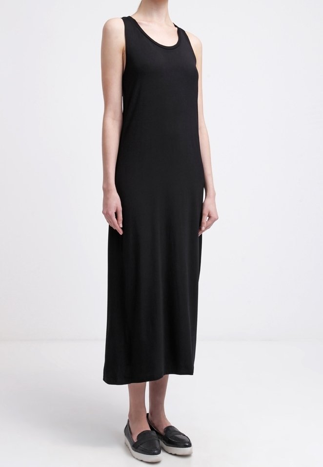 Robe midi noire sans manches à encolure dégagée portée par une personne debout, associée à des chaussures noires sans lacets à semelles blanches.