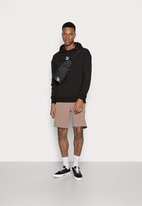 Felpa nera con tasca a marsupio, pantaloni beige e sneaker nere. Una piccola borsa a tracolla nera è indossata sul corpo. Design minimalista.