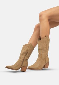 Botas de cowboy de ante marrón con punta afilada, adornos de costura en forma de zigzag, cremalleras laterales y tacones bloque apilados, posicionadas en un ángulo.