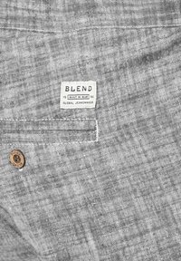 Sivi materijal s teksturiranim uzorkom. Ima tkanu oznaku "BLEND BUILT IN BLUE" i drveni gumb na džepu.