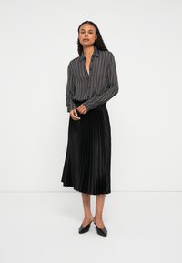 Vrouw in een langemouwen blouse met verticale strepen en een zwarte geplooide midi-rok, staande tegen een effen witte achtergrond.