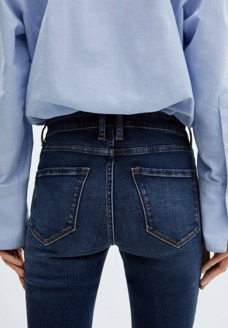 Jeans en denim foncé avec une texture lisse, taille haute, deux poches arrière et des coutures orange, associés à un haut à manches longues bleu clair.