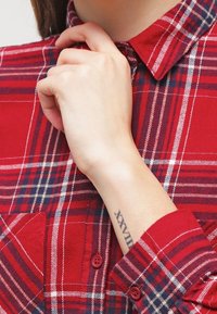 Red plaid button-up shirt with white and blue accents, featuring a classic collar, chest pocket, and rolled-up sleeves. Visible tattoo on wrist.