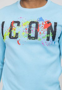 Sweatshirt bleu clair avec de grandes lettres noires épelant "ICON", agrémenté d'éclaboussures colorées en rouge, vert, bleu et rose. Matériel en coton doux.