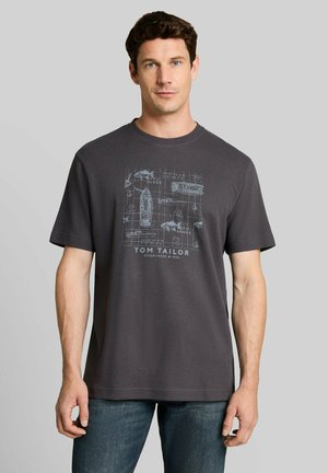 Mann trägt dunkelgraues kurzärmeliges T-Shirt mit Boots- und Fisch-Design und dem Text "Tom Tailor", kombiniert mit blauen Jeans, steht vor einem einfarbigen Hintergrund.
