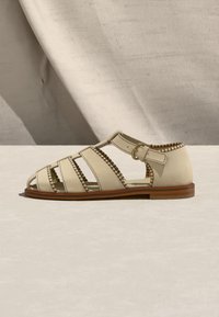 Μπεζ επίπεδη δερμάτινη σάνδαλα με πολλαπλούς ιμάντες, πλαϊνή αγκράφα, ραμμένες λεπτομέρειες κατά μήκος των άκρων και ξύλινη σόλα.