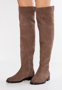 Brązowe zamszowe kozaki za kolano z płaskim obcasem i bocznym zamkiem. Miękka tekstura, bezszwowy design i zaokrąglony kształt czubka.