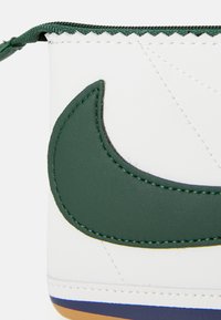 Hvid læderprodukt med et stort grønt swoosh-logo, detaljeret syning og lynlåsafslutning, samt en multicolorer sålakkent.
