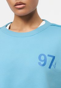 Gros plan sur une personne portant un haut bleu clair avec un sous-shirt blanc, portant le numéro "97" et le mot « Lover » imprimés sur la poitrine.