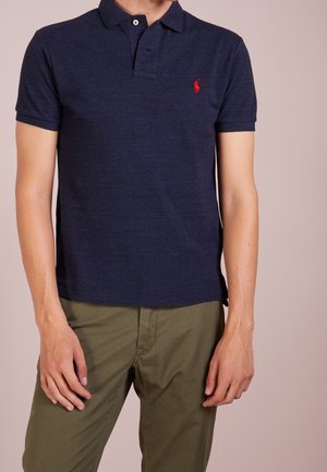 Man draagt een navyblauw poloshirt met een rood logo en olijfgroene broek, staand tegen een licht beige achtergrond.