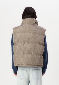Šedá prošívaná vesta s vysokým límcem a průramky, nošená přes modrou košili s dlouhým rukávem. Má volný střih a prošívaný design.