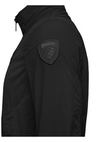 Mustat takki, jossa on korkea kaulus, teksturoitu kangas ja matta musta logo-patch olkapäällä. Varustettu vetoketjulla edessä.