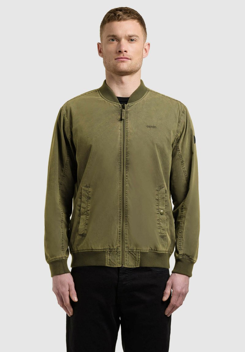 Homme debout face à l'avant portant une veste aviateur vert olive avec fermeture éclair et un pantalon noir sur un fond uni.