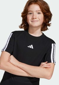 T-shirt noir à manches courtes avec des rayures blanches sur les épaules et un logo blanc sur la poitrine. Le tissu semble lisse et extensible.