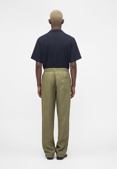 Άνδρας με κοντά ξανθά μαλλιά φοράει μαύρο κοντομάνικο πουκάμισο μπουζωμένο σε φαρδιά ελιάς παντελόνια, φαίνεται από πίσω σε απλό φόντο.