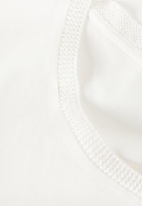Tissu blanc avec une texture lisse, présentant une bordure perforée le long du col, mettant en valeur une forme arrondie et des bords nets.