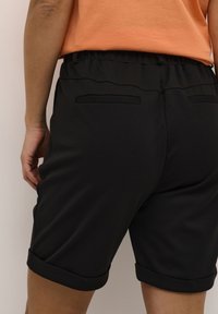 Mustat lyhyet housut, joissa on sileä rakenne, joustava vyötärö ja käänteet. Kaksi takataskua on näkyvissä. Mallilla on päällään oranssi paita.