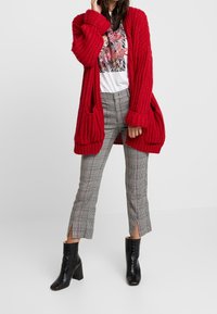 Cárdigan de punto rojo con ajuste holgado y textura acanalada, combinado con pantalones a cuadros con aberturas laterales y botas de tacón negras.