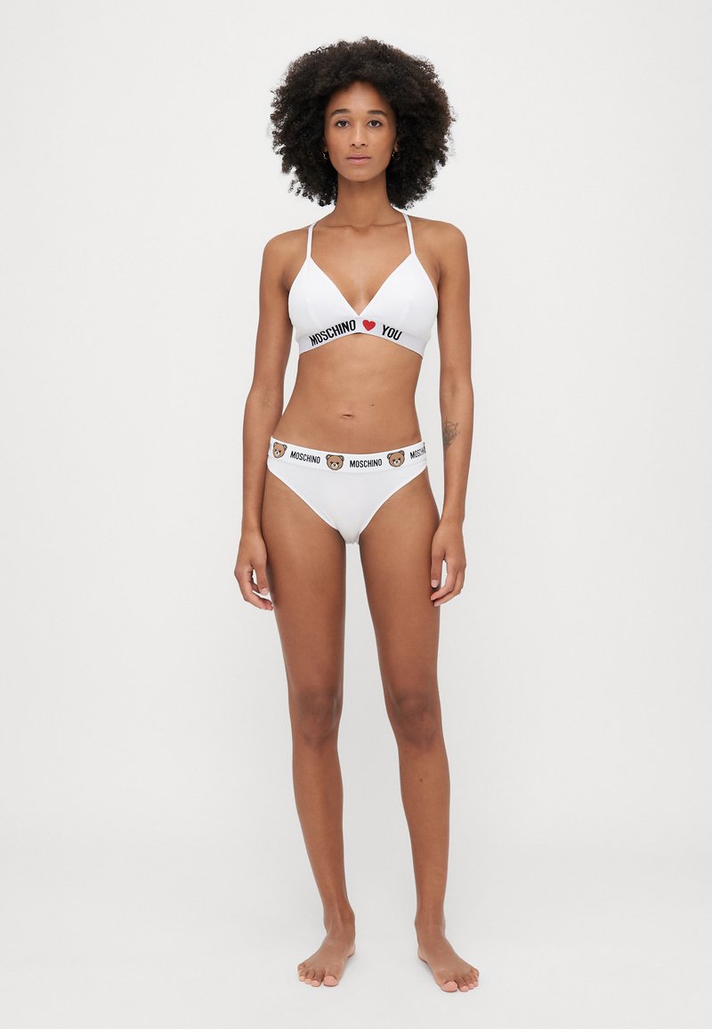 Balts bikini komplekts ar trīsstūra formu krūšturi, uz kura ir melns "MOSCHINO I ❤️ YOU" teksts, un augsto jostasvietu biksītes ar lāča logotipa akcentu.