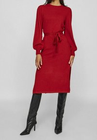 Czerwona dzianinowa sukienka z długimi rękawami, wiązaniem w talii, bocznymi rozcięciami i długością do połowy łydki. Zestawiona z dopasowanymi czarnymi kozakami za kolano.