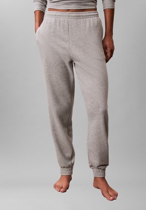 Persona che indossa pantaloni della tuta grigio chiaro con polsini e vita elasticizzati, in piedi a piedi nudi contro uno sfondo grigio uniforme.