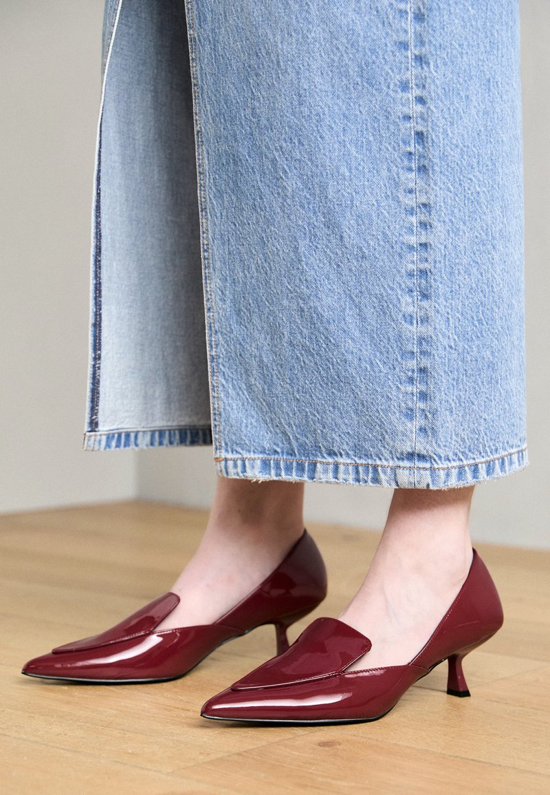 Talons en cuir verni rouge avec un bout pointu, présentant un design découpé et un talon stiletto bas, portés avec une jupe midi en denim.
