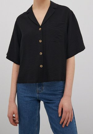 Persona que lleva una camisa negra de manga corta con botones y bolsillo en el pecho, y vaqueros azules de mezclilla sobre un fondo liso.