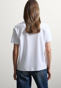 Camiseta blanca de algodón con mangas cortas, corte holgado, escote redondo y diseño sin costuras, combinada con pantalones vaqueros azules.