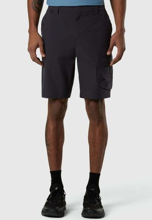 Persona che indossa pantaloncini cargo neri fino al ginocchio, scarpe da ginnastica nere e calzini neri, in piedi davanti a uno sfondo semplice.