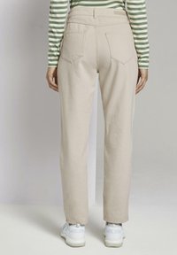 Pantalons beige taille haute avec une jambe droite, deux poches arrière et une texture lisse. Portés avec des baskets blanches et une chemise rayée.