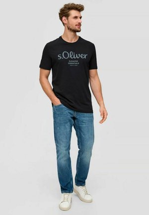 Mann trägt ein schwarzes T-Shirt mit s.Oliver-Logo, blaue Jeans und weiße Sneaker, steht mit einer Hand in der Tasche vor einem schlichten Hintergrund.