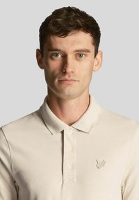 Beige Poloshirt aus strukturiertem Stoff, mit Kragen und einer dreiknöpfigen Knopfleiste, mit einem gestickten Logo auf der linken Brust.