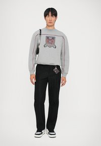 Felpa grigia con logo a scudo, accenti rossi, abbinata a pantaloni neri texturizzati. Dettagli notevoli includono una piccola borsa nera e un portachiavi.