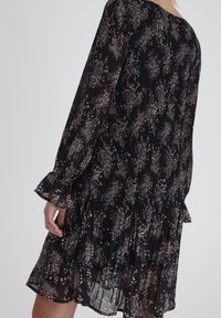 Svart blommönstrad plisserad klänning med långa ärmar, volangärmar och flerlagad nederdel. Blommönster i vitt, rosa och blått på genomskinligt tyg.