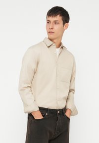 Μπεζ πουκάμισο με γιακά, μία τσέπη στο στήθος και γυρισμένα μανίκια, συνδυασμένο με σκούρα τζιν σε ενιαία απόχρωση.