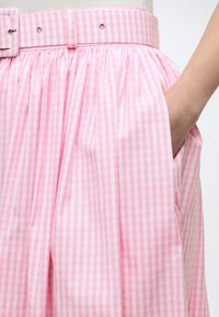 Ροζ και λευκή καρό φούστα με ταιριαστή ζώνη και χέρι τοποθετημένο μέσα στην πλαϊνή τσέπη, παρουσιασμένη σε ένα άτομο με φόντο έναν ανοιχτόχρωμο χώρο.