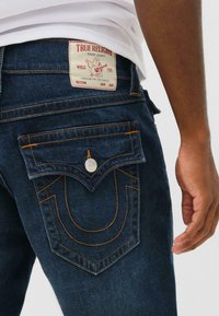 Donkerblauwe spijkerbroek met oranje stiksels, met een achterkant pocketontwerp en een merklabel met een gitaarafbeelding.