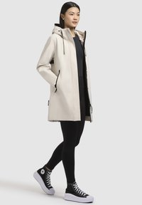 Manteau de pluie beige imperméable avec capuche, doté de cordons de serrage réglables, d'accents noirs, de poches latérales et d'une coupe droite. Associe avec des leggings noirs et des baskets montantes.