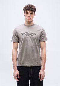 Νέος άνδρας με σγουρά μαλλιά, που φοράει γκρι μπλουζάκι με λογότυπο Aston Martin και σκούρο τζιν, στέκεται μπροστά σε απλό ανοιχτό φόντο.