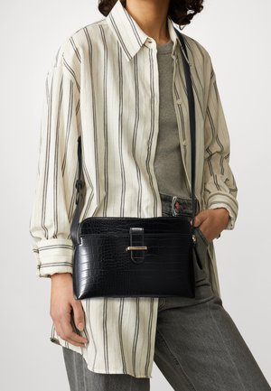 Femme portant une chemise à rayures crème sur un haut gris et un jean, avec un sac bandoulière en cuir embossé crocodile noir, orné d'une boucle dorée.