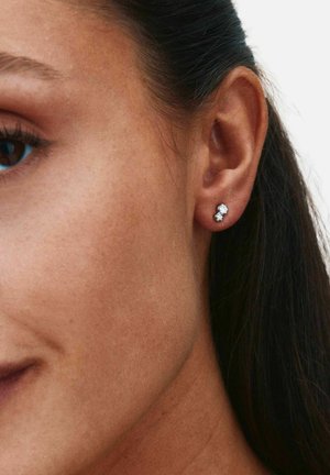 Orecchio destro di donna che indossa piccoli orecchini a lobo con doppia pietra, con capelli scuri raccolti dietro l'orecchio e pelle liscia visibile.