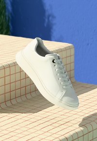 Baskets blanches en cuir avec une finition lisse, un design à lacets et une semelle en caoutchouc texturée, posées sur une surface à motif de grille.