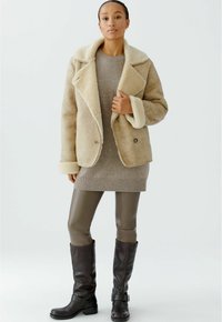 Femme portant une veste en shearling beige par-dessus une robe en maille beige, des leggings en cuir marron et des bottes hautes marron foncé, se tenant contre un fond blanc.