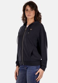 Felpa nera con zip e dettagli in oro, cappuccio con coulisse e tasche laterali. Realizzata in tessuto morbido con una vestibilità rilassata e polsini a coste.