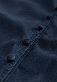 Tissu en denim bleu marine avec une surface texturée, présentant des boutons ronds recouverts de tissu le long du centre devant et des détails de couture visibles.