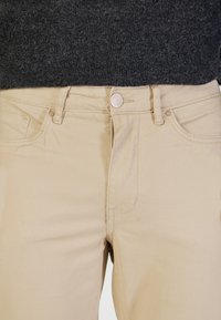 Pantalones de algodón beige con cinco bolsillos, cremallera frontal y cierre con botón de metal; textura suave y diseño casual.