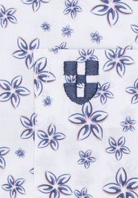 Biała tkanina z niebieskim kwiatowym wzorem, z kieszenią z niebieskim haftowanym emblematem przedstawiającym wzór tarczy.